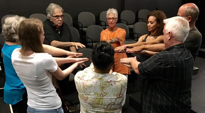 A group of improv students in a circle with eyes closed and arms outstretched.